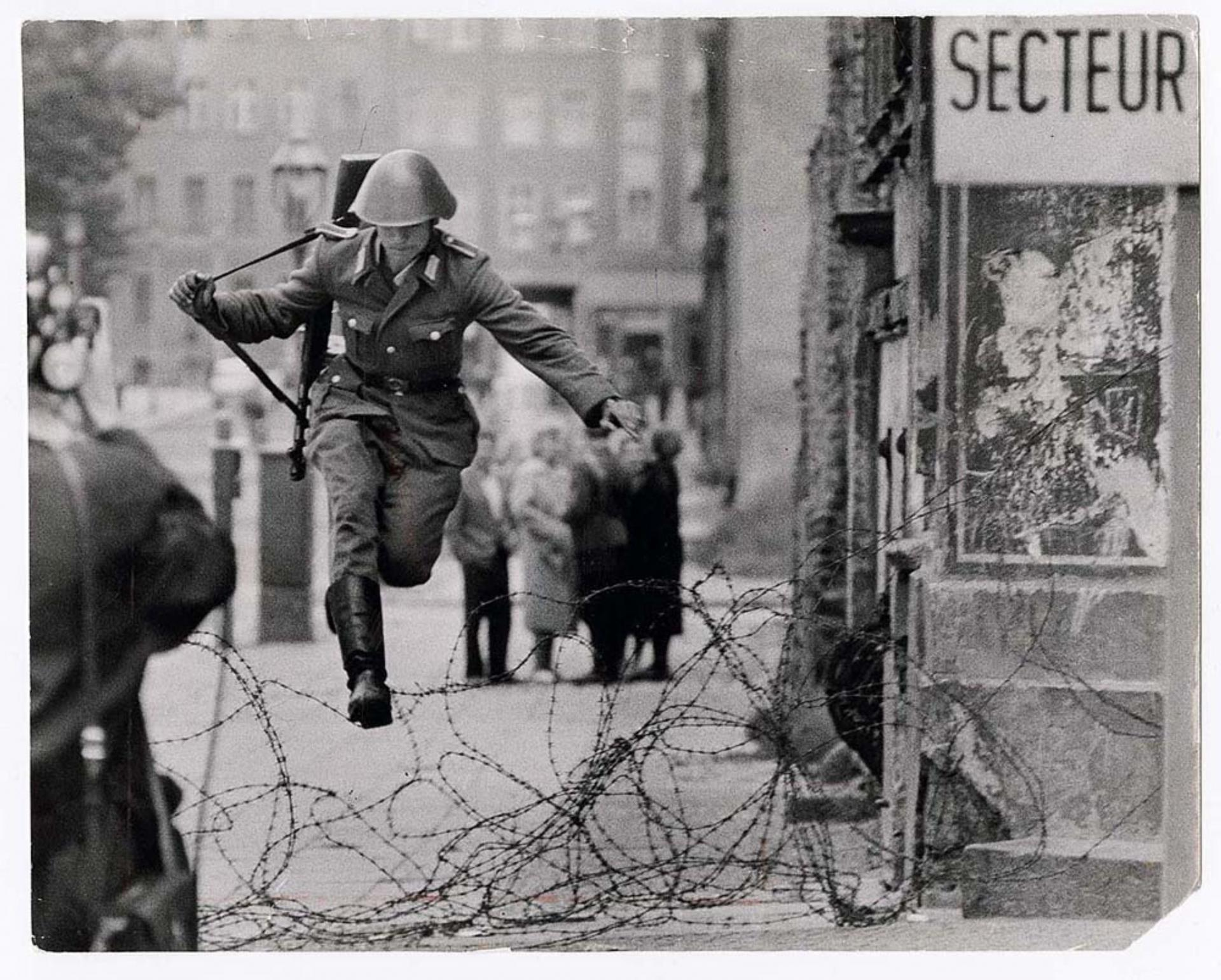Photo en noir et blanc d’un soldat en uniforme qui saute au dessus de barbelés.